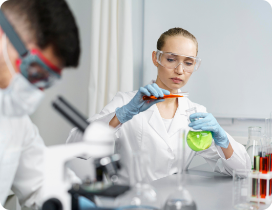Pair of scientists. The woman transfers a liquid from one container to the other, while the man analyzes something under his microscope.